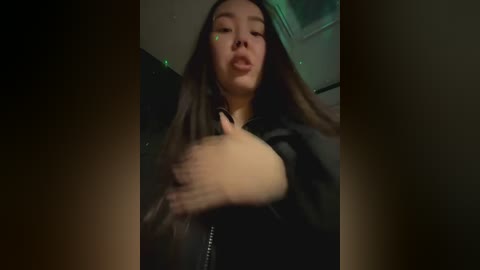 Media: A video of a young woman with long, dark hair and fair skin, wearing a black jacket, standing in a dimly lit room with green lighting. She has a neutral expression and her hands are partially raised.