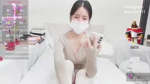 Media: Video of a young East Asian woman with long black hair, wearing a beige dress, white face mask, and holding a phone. Background includes a bed with white linens, a desk, and a partially visible TV screen displaying a game.