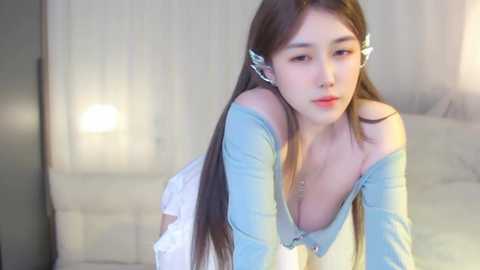 Media: Video of an Asian woman with long brown hair in a light blue off-shoulder top, leaning forward on a bed with white sheets. Soft lighting and blurred background.