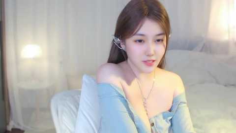 Media: A video of an Asian woman with fair skin and long brown hair, wearing a light blue off-shoulder top, sitting on a bed in a softly lit bedroom.