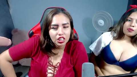 Media: Video of two women: one with long brown hair in a red shirt, and another in a blue bra. They're in a room with a fan, red gaming chair, and a blue wall.