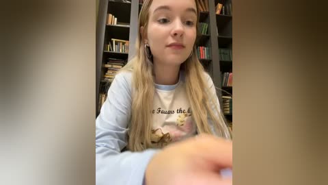 Media: Video of a young blonde girl in a light blue sweatshirt reading \"The Fault in Our Stars\" indoors, surrounded by bookshelves filled with books.