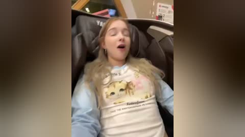 Media: Video of a young, fair-skinned blonde woman with a relaxed expression, wearing a white t-shirt with an animal design, reclining in a dark leather chair. Background includes a bookshelf and a window with a view of a green tree.