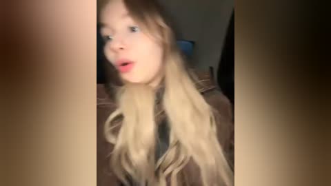 Media: A blurry, candid video of a fair-skinned woman with long, wavy blonde hair, wearing a brown jacket, captured indoors with a dim, warm lighting.