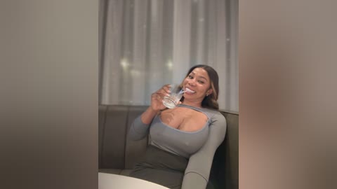 Media: Video of a smiling, curvy woman with medium skin tone, long dark hair, wearing a tight, low-cut gray top, drinking water, sitting on a gray couch in a modern, dimly lit room.