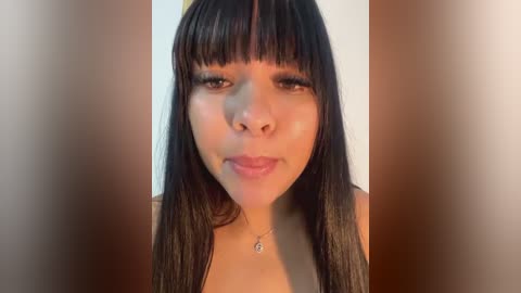 Media: Video of a young woman with medium skin tone, straight black hair with bangs, wearing a silver necklace. She has a neutral expression, with a plain, light-colored background.