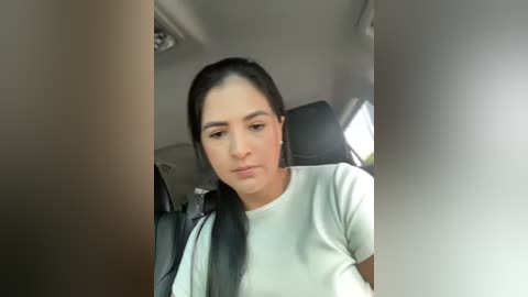 Media: Video of a young woman with long black hair, fair skin, and a neutral expression, wearing a white t-shirt, sitting in a car with black seats and a gray headrest.