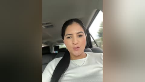 Media: A video of a woman with light brown skin and dark hair tied back, wearing a white shirt, driving a car. The car's interior is visible with a gray seat and window.