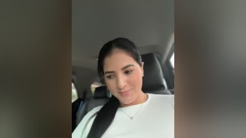 Media: Video of a Latina woman with long black hair, wearing a white t-shirt, seated in a car with a black seatbelt, looking down, blurred background.