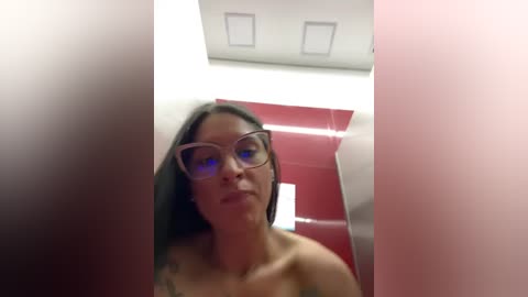 Media: Video of a woman with dark hair, glasses, and a tattoo on her shoulder, standing in a red-tiled bathroom with a white ceiling and rectangular lights.