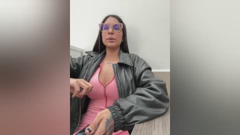 Media: A video of a woman with long black hair, wearing glasses, a pink top, and a black leather jacket, seated on a chair.