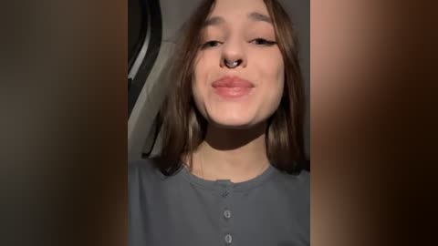 Media: Video of a young woman with fair skin and straight brown hair, wearing a gray shirt. She has a septum ring and a closed-mouth smile. Background is blurred, possibly an indoor setting.