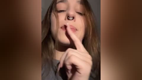 Media: Video of a young woman with light skin, long brown hair, and a septum piercing. She's wearing a gray shirt and is holding a finger to her lips, suggesting shushing.