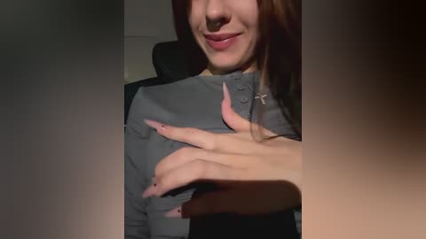 Media: Video of a smiling, fair-skinned woman with straight brown hair, wearing a grey button-up shirt. Her left hand, with long pink nails, rests on her chest. She sits on a black chair in a dimly lit room.