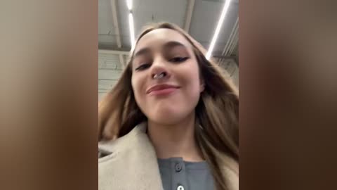 Media: A young woman with light skin, long brown hair, and a nose ring smiles, wearing a beige coat over a gray top, in a dimly lit room with exposed brick walls and fluorescent lights.