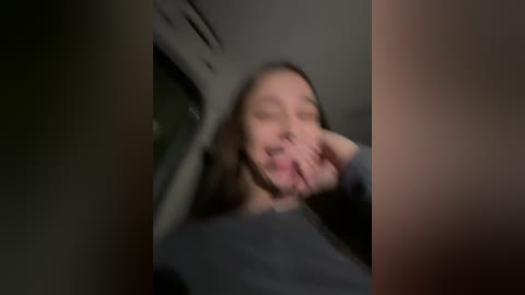 Media: A blurred video of a woman with long brown hair, wearing a dark shirt, covering her mouth with her right hand, inside a car.