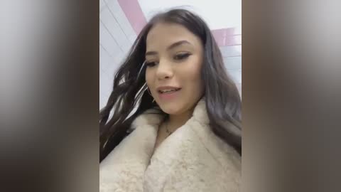 Media: A close-up video of a young woman with long, dark hair, wearing large hoop earrings and a beige fur coat, smiling slightly, in a bathroom with white and pink tiled walls.