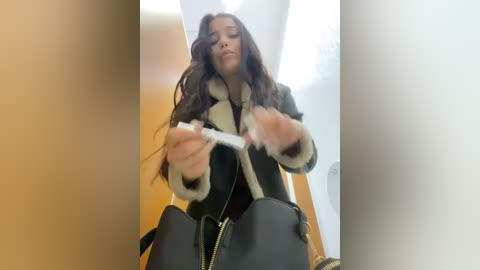 Media: Video of a young woman with long, wavy brown hair, wearing a black leather jacket with fur trim, holding a syringe, seated in a dimly lit room with beige walls and a wooden door.