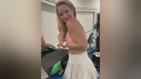 Media: Video of a blonde woman with fair skin, wearing a pink sports bra and white pleated skirt, smiling in a gym with workout equipment and mats.