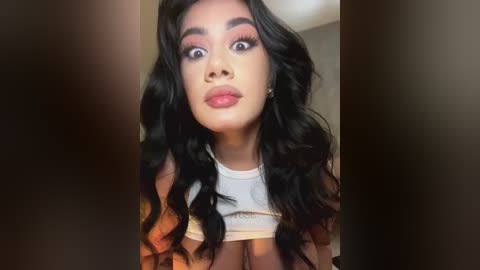 Media: Video of a young Latina woman with long, wavy black hair, wearing a white crop top, standing indoors with a surprised expression.