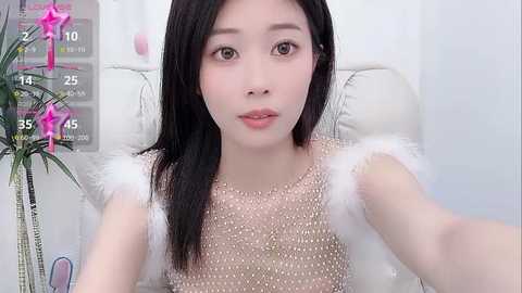 Media: Video of an East Asian woman with long black hair, fair skin, wearing a white fur-trimmed, beaded top, sitting on a white leather chair. Background features a pink star filter and a potted plant.