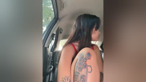 Media: Video of a woman with long dark hair, wearing a red top, sitting in a car. She has a large colorful tattoo on her left shoulder depicting a flower and butterfly. The interior of the car is visible.