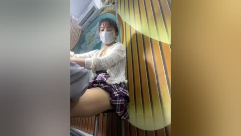 Media: Video of a young Asian woman wearing a white face mask, plaid skirt, and sweater, seated in a wooden chair with a green and yellow background.