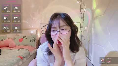 Media: A video of a young Asian woman with long black hair, wearing glasses, lying on a bed, holding a phone to her mouth, in a cozy bedroom with a pink wall.