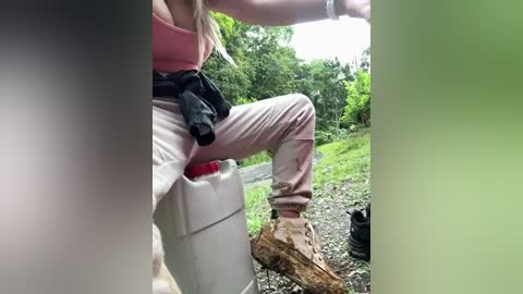 Media: Video of a woman in a pink tank top and beige pants, sitting on a white cooler, with a green background featuring trees and grass, wearing brown hiking boots and a blue watch.