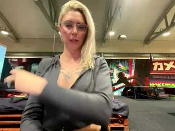 Media: Video of a blonde, fair-skinned woman in glasses, wearing a gray hoodie, adjusting her hair in a gym with colorful motivational posters and workout equipment in the background.