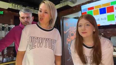 Media: Video of three young women, one blonde and two with red hair, wearing white New York T-shirts, standing in a dimly lit bar. Background features a bartender and a colorful menu board.