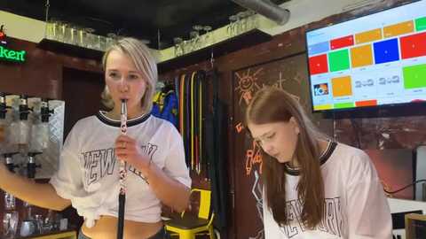 Media: Video of two blonde women in white \"MEN\" T-shirts playing ukuleles in a dimly lit bar. Background features a colorful digital screen, bottles, and a glass rack.