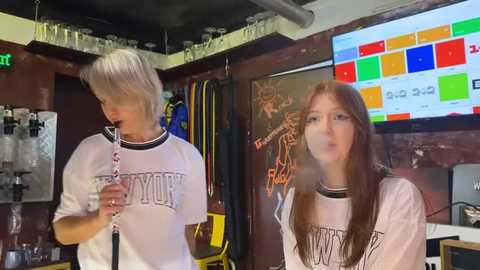 Media: Video of a young woman with long, straight brown hair and a white T-shirt, playing a clarinet in a cozy room with a colorful mosaic TV screen and a cluttered background.