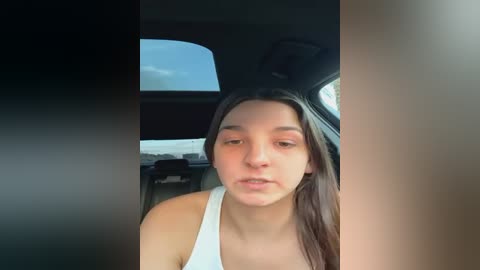 Media: Video of a young woman with long, straight brown hair, wearing a white tank top, sitting in a car with a view of a blue sky through the window.