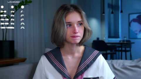 Media: Video of a young Caucasian woman with light brown hair, wearing a white sailor collar shirt, seated indoors. Background features modern decor with a plant, a table, and abstract artwork.