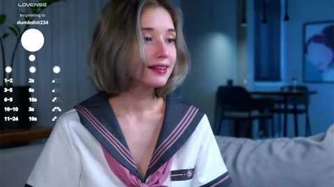 Media: Video of a young woman with shoulder-length blonde hair, wearing a white sailor-style top with pink accents, smiling softly in a dimly lit living room.