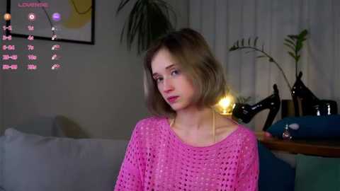 Media: Video of a Caucasian woman with light skin, short blonde hair, wearing a pink crochet top, sitting on a sofa, indoors, dim lighting, plant in the background.