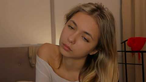 Media: Video of a young Caucasian woman with long, blonde hair, wearing a white off-shoulder top, leaning slightly forward with a contemplative expression. Background includes a beige wall, a black metal shelf, and a red garment hanging on it.