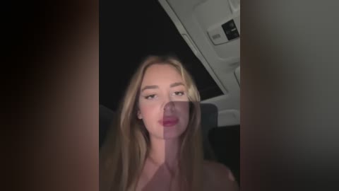 Media: Video of a blonde woman with long hair, wearing makeup, sitting in a car with a dark interior. Her lips are covered by a see-through plastic covering.