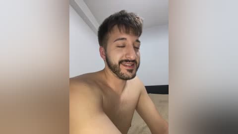 Media: Video of a young, fair-skinned, bearded man with short brown hair, smiling, topless, sitting indoors, with blurred white walls and beige carpet background.