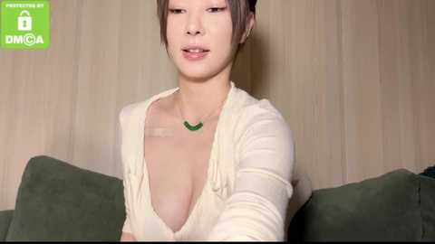 Media: Video of an East Asian woman with short brown hair, light skin, wearing a low-cut cream top revealing cleavage, green necklace, seated on a green couch against wooden-paneled wall, with \"PROMISING OF THAIWIFE\" watermark in the top left.