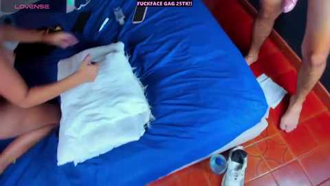 Media: Video of a man's bare feet and legs, wearing a blue shirt, standing next to a bed with a torn white pillow on a blue bedspread. Text at top reads \"LOVENSB,\" and at bottom reads \"FUCKGAGSTREET.\