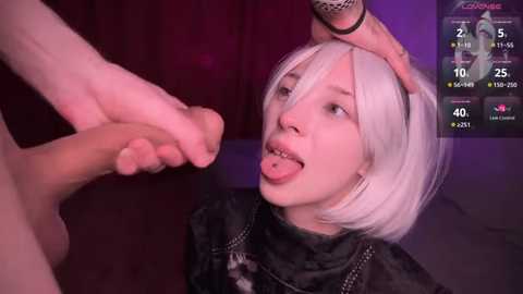 Media: Video of a white woman with platinum blonde bob haircut and a black top, tongue sticking out, being held by a man's hand, in a dimly lit room with purple lighting.