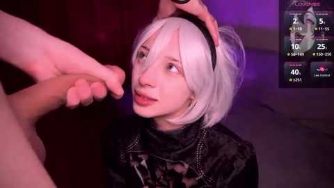 Media: Video of a pale-skinned woman with white bob haircut and black headband, wearing a black velvet outfit, being caressed by a hand in a dimly lit room.