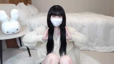 Media: Video of a young woman with long black hair, wearing a white face mask, white robe, and sitting on a fluffy white rug. She has a serene expression. Behind her, a white bed with white pillows and a white stuffed bunny on a side table.