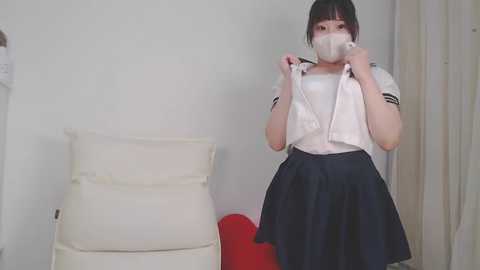 Media: Video of an Asian woman with short black hair, wearing a white sailor blouse, black skirt, and face mask, adjusting her mask, standing in a minimalist, white-walled room with a beige chair and red cushion.