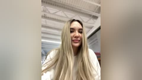 Media: A video of a young woman with long, straight blonde hair, wearing a white sweater, standing in a modern room with white ceiling beams and a blurred background.