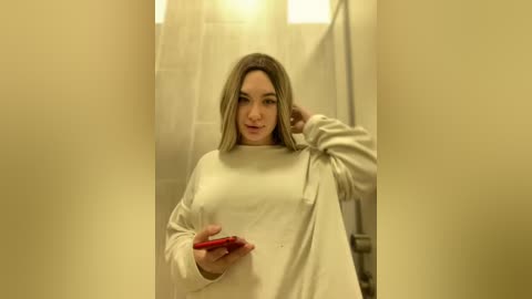 Media: Video of a young woman with shoulder-length blonde hair, wearing a loose, off-white sweater, holding a red smartphone, standing in a beige-tiled bathroom with a shower curtain.