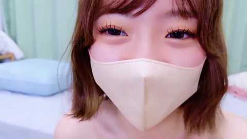 Media: Video of an Asian woman with light skin, shoulder-length brown hair, and brown eyes. She wears a white medical mask and a light blue hospital gown, sitting on a bed with a white pillow and a pale green wall in the background.