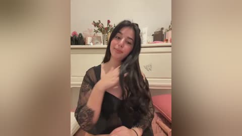 Media: Video of a young Asian woman with long black hair, wearing a sheer black lace top, sitting on a bed in a bedroom with a white dresser, floral decorations, and pink pillow.